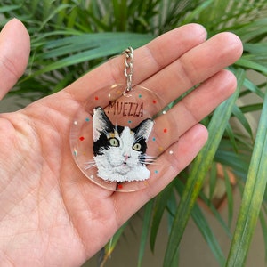 May include: A clear, round keychain featuring a hand-painted portrait of a cat with black, white, and yellow markings. The cat's name, "MUEZZA," is written above the portrait. The keychain has a silver chain and is decorated with colorful dots.