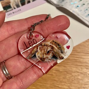 May include: Heart-shaped keychain with a painted image of a brown and tan bunny. The keychain has the name "Leo" written on it and small pink hearts. A silver keyring is attached to the top.