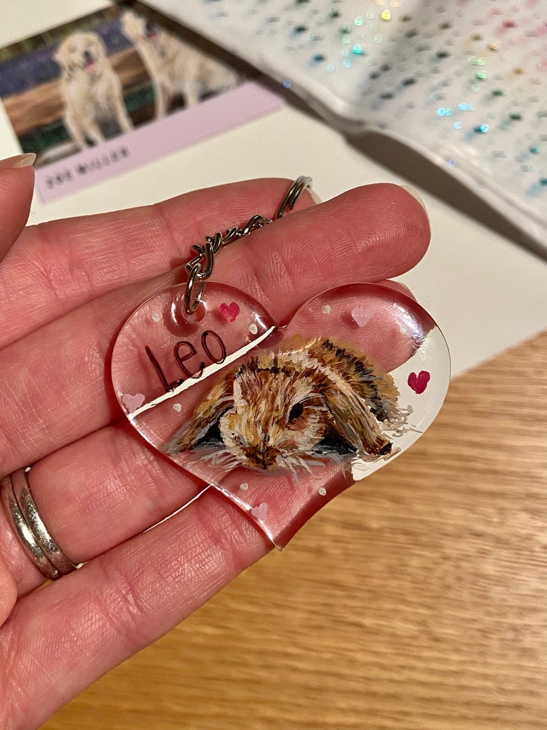 May include: Heart-shaped keychain with a painted image of a brown and tan bunny. The keychain has the name "Leo" written on it and small pink hearts. A silver keyring is attached to the top.