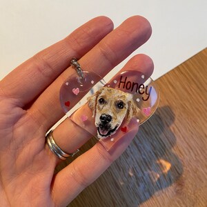 May include: A clear, heart-shaped keychain featuring a painted portrait of a golden retriever dog. The dog's name, "Honey," is written in black script. Small pink hearts and white dots decorate the keychain, which is attached to a silver keyring.