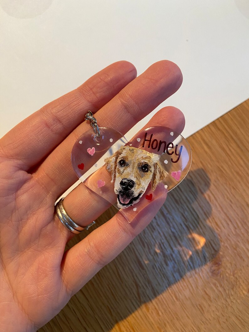 May include: A clear, heart-shaped keychain featuring a painted portrait of a golden retriever dog. The dog's name, "Honey," is written in black script. Small pink hearts and white dots decorate the keychain, which is attached to a silver keyring.