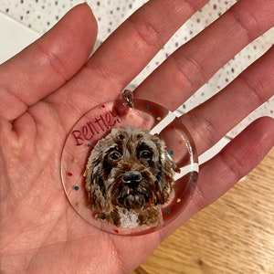 May include: A clear, round pendant featuring a painted portrait of a brown and white dog with the name "Bentley" in red. The pendant has a silver loop and is decorated with colorful confetti.