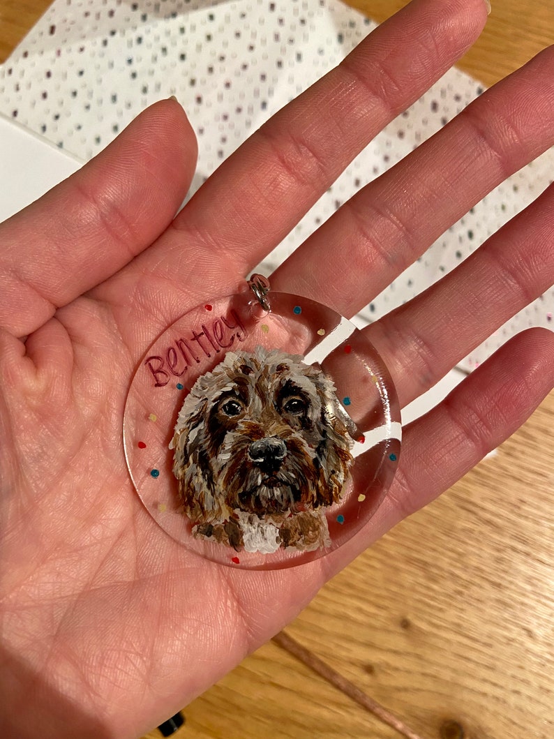 May include: A clear, round pendant featuring a painted portrait of a brown and white dog with the name "Bentley" in red. The pendant has a silver loop and is decorated with colorful confetti.