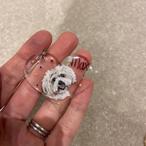 May include: A clear, heart-shaped keychain featuring a painted portrait of a white dog with the name "Max" in black lettering. The keychain has a silver ring and is decorated with colorful dots.