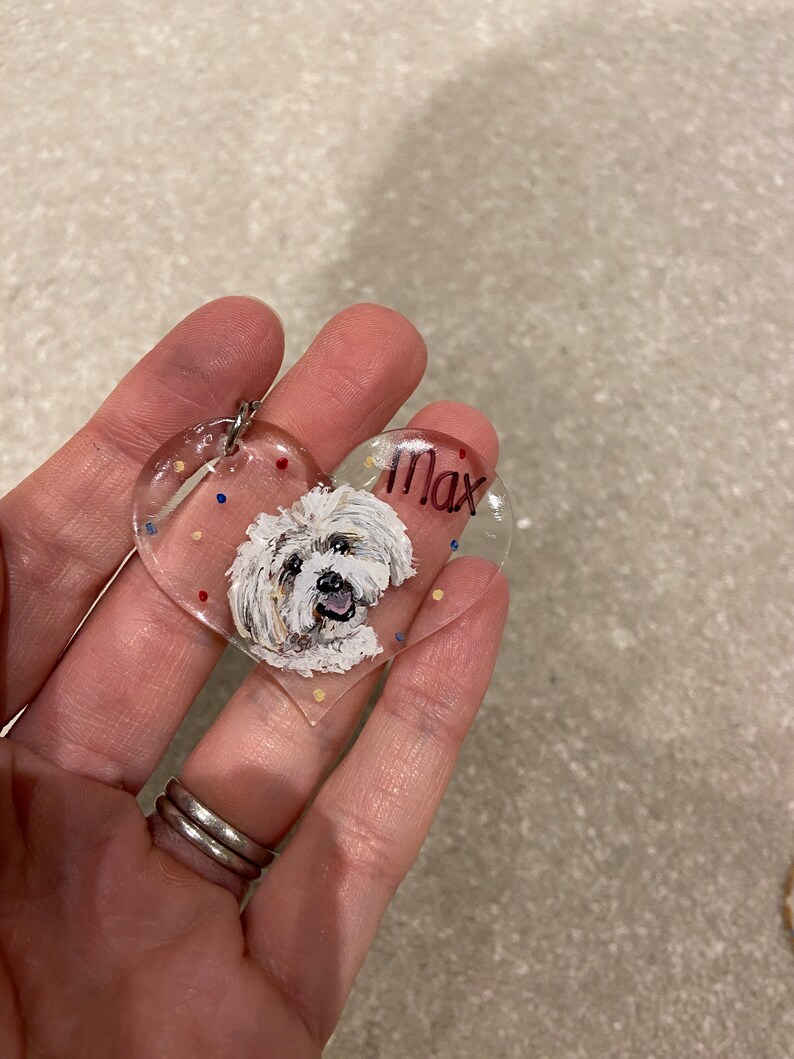 May include: A clear, heart-shaped keychain featuring a painted portrait of a white dog with the name "Max" in black lettering. The keychain has a silver ring and is decorated with colorful dots.