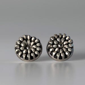 Könnte beinhalten: Ein Paar silberne Ohrstecker mit einem runden Design, das ein erhabenes, strukturiertes, blumenartiges Muster aufweist.