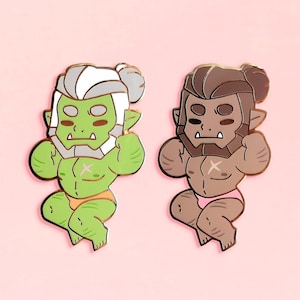 Puede incluir: Dos pines de esmalte con representaciones de dibujos animados de orcos verdes y marrones. El orco verde tiene el pelo y la barba blancos, mientras que el orco marrón tiene el pelo marrón. Ambos orcos llevan ropa interior rosa y tienen una cruz en el pecho.