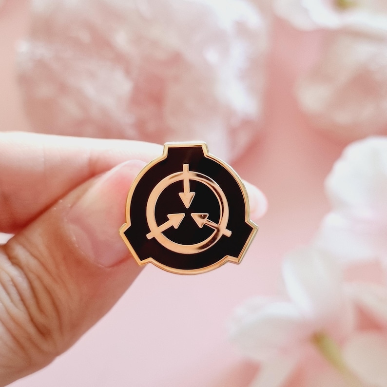 May include: A gold-colored pin with a black enamel design. The design is a stylized symbol with three arrows pointing inwards, surrounded by a circle. The symbol is often associated with the fictional organization known as the SCP Foundation.