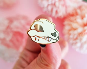 May include: Gold enamel pin featuring a white skull with a heart-shaped eye and a small sparkle design. The skull has a toothy grin.