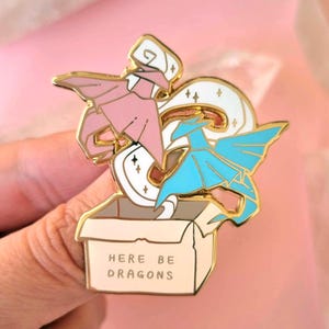 May include: Gold enamel pin featuring two origami dragons, one pink and one blue, emerging from a box with the text "Here Be Dragons".