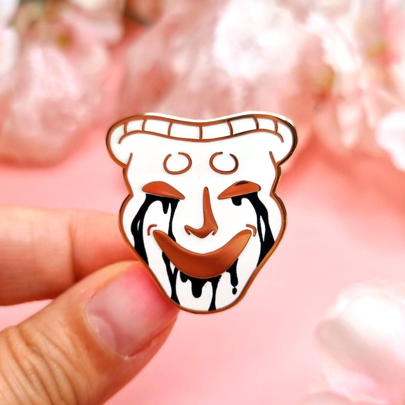 May include: A white enamel pin in the shape of a smiling mask with black tears and a gold outline. The mask has a brown nose and mouth, with two small circles above the eyes. The pin is held in a hand against a pink background.