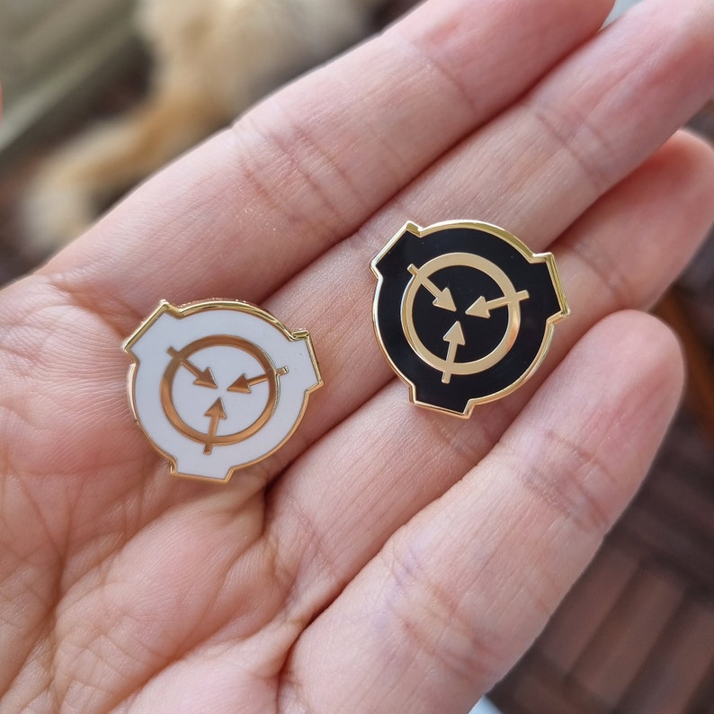 May include: Two gold-colored metal pins with a black and white circular symbol. The symbol is a circle with three arrows pointing inward. The pins are on a hand.