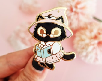May include: A black cat enamel pin wearing a pink witch hat and dress, with gold accents. The cat is holding a light blue book with gold stars. The pin has a gold outline and is set against a soft pink background.