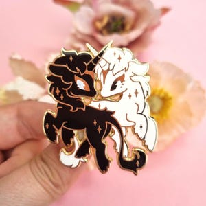 May include: A gold-toned metal enamel pin featuring two intertwined unicorn figures, one solid black and one solid white, both decorated with small gold star accents and gold outlines, creating a contrasting yin and yang style design.