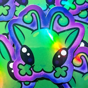 May include: A collection of stickers featuring a cartoon alien design. The stickers have a green body with a holographic sheen, and are outlined in purple and blue. The alien has large black eyes and a small mouth.