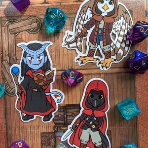 May include: Three illustrated stickers featuring fantasy characters. The first sticker is a blue-skinned humanoid with horns, wearing a black robe and holding a staff and a book. The second sticker is a bird-like creature with brown and white feathers, wearing a brown hooded cloak and holding a bow and arrow. The third sticker is a black bird-like creature with red eyes, wearing a red hooded cloak and holding a dagger.