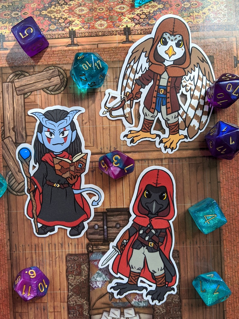 May include: Three illustrated stickers featuring fantasy characters. The first sticker is a blue-skinned humanoid with horns, wearing a black robe and holding a staff and a book. The second sticker is a bird-like creature with brown and white feathers, wearing a brown hooded cloak and holding a bow and arrow. The third sticker is a black bird-like creature with red eyes, wearing a red hooded cloak and holding a dagger.