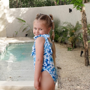 May include: A young girl wearing a blue and white floral print swimsuit stands near a pool. The one-piece swimsuit has a ruffled neckline and a botanical pattern. The girl has blonde hair styled in pigtails.