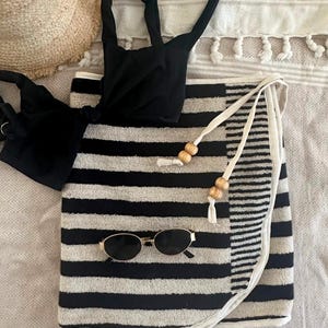 May include: A black bikini top, a black and white striped wrap skirt with white ties and wooden beads, a pair of oval sunglasses with gold frames, and a straw hat. The wrap skirt features horizontal stripes and a white trim along the edge.