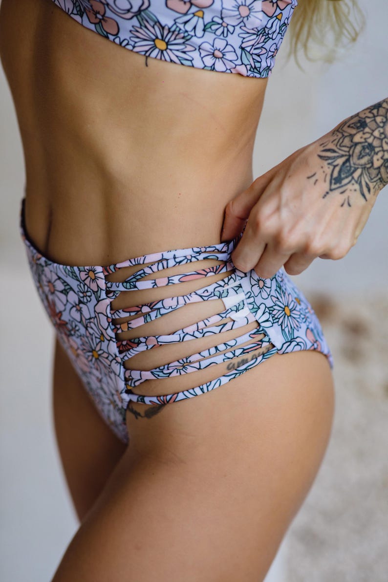 K&ouml;nnte beinhalten: High-Waist-Bikinihose mit hellblauem Hintergrund und einem sich wiederholenden Blumenmuster aus wei&szlig;en, rosafarbenen und dunkelblauen G&auml;nsebl&uuml;mchen. Die Seiten des Kleidungsst&uuml;cks verf&uuml;gen &uuml;ber eine Reihe horizontaler Cut-Out-Riemen.