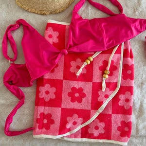 May include: A bright pink bikini top with thin straps and a matching wrap skirt featuring a pink and red checkered pattern with floral motifs, white trim, and wooden beads on the ties, accompanied by a tan straw hat with a frayed edge.