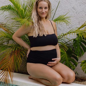 K&ouml;nnte beinhalten: Ein zweiteiliger schwarzer Badeanzug bestehend aus einem Bandeau-Oberteil mit d&uuml;nnen Tr&auml;gern und einer hoch taillierten Hose mit gerafften Seitendetails. Die Badebekleidung besteht aus einfarbigem schwarzem Stoff und bedeckt den Oberk&ouml;rper.