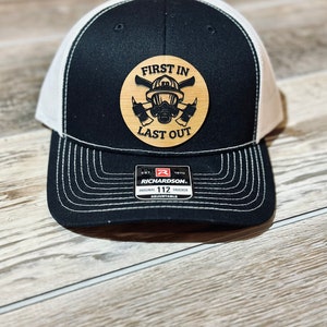 May include: Black and white trucker hat with a wood patch that says "First In Last Out" with a firefighter helmet and crossed axes design.