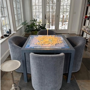 May include: A square, dark gray table with a board game in progress, surrounded by four gray upholstered barrel chairs. A small lamp illuminates the game. A small, silver side table is in the foreground.