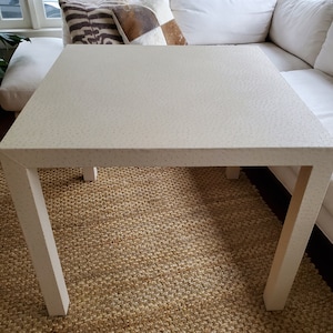 May include: A white square coffee table with a speckled surface and four legs. The table is sitting on a woven rug.