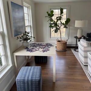 May include: A white table with a partially completed jigsaw puzzle on top. A blue and white patterned ottoman sits in front of the table. A large potted fiddle leaf fig plant is in the background.