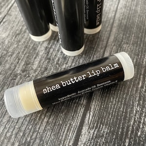 May include: Close-up of shea butter lip balm in a black tube with white text. The label reads "shea butter lip balm" and lists ingredients. Several other tubes are visible in the background.