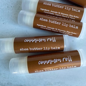 May include: Close-up of several shea butter lip balm tubes. The tubes have a brown label with white text that reads "shea butter lip balm" and "cinnamon roll". Ingredients include avocado oil, beeswax, shea butter, cocoa butter, and organic vitamin E.