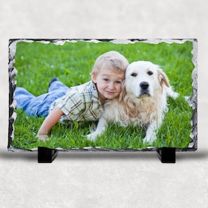 May include: A young boy with blonde hair is lying on the grass with a golden retriever dog. The boy is wearing a plaid shirt and blue jeans. The dog is looking at the camera.