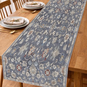 Op de afbeelding: Een rechthoekige, blauwe tafelloper met een ingewikkeld bloemmotief in grijstinten, rood en beige. De loper ligt op een houten tafel en voegt een decoratief element toe aan de eetruimte. Afmetingen van de loper niet zichtbaar.