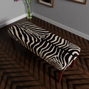 以下が含まれることがあります： 黒とクリーム色のゼブラ柄の長方形ベンチ。 ベンチには4本の木製の脚があり、濃い茶色の木製の床に置かれています。 ベンチの長さは約122cmです。