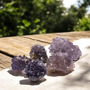 May include: Five amethyst crystal clusters in varying sizes and shades of purple are arranged on a white cloth. The crystals have a natural, rough texture and are illuminated by sunlight. The background features a wooden surface and blurred green foliage.