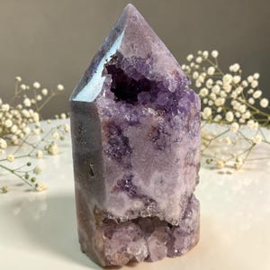 May include: A large, amethyst crystal point with a polished, faceted top and a rough, textured body. The crystal features a deep purple hue with lighter lavender and white inclusions. The interior reveals a geode-like cavity filled with sparkling crystals.