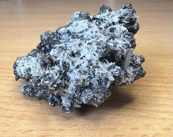 May include: A natural mineral specimen featuring a cluster of dark gray, metallic-looking crystals with a coating of clear, white quartz crystals. The rough, textured surface of the mineral is displayed on a light brown wooden surface.