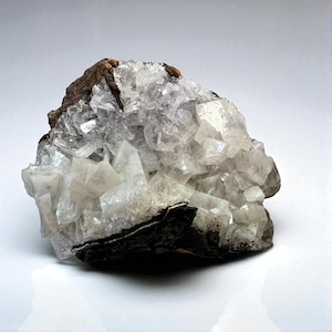 May include: A natural, raw crystal cluster with a mix of clear and opaque white crystalline structures. The base of the cluster is dark gray and brown, with a rough, textured appearance. The crystals have a sparkling, reflective quality.