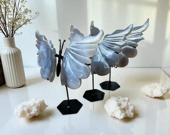 May include: Three decorative butterfly sculptures with gray and white patterned wings are displayed on black stands. The butterfly wings are made of a translucent material. White, coral-like objects are scattered around the butterflies.