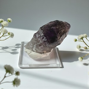 May include: A large, raw amethyst crystal with a deep purple hue, mounted on a clear acrylic stand. The crystal is partially opaque and has a rough, textured surface. White flowers are arranged around the crystal, adding a delicate contrast.