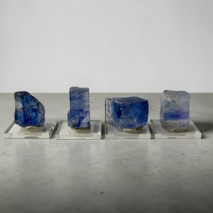 May include: Four blue crystals of varying shapes are displayed on clear acrylic stands. The crystals have a deep blue color with some areas appearing translucent. The stands are square and elevate the crystals, showcasing their natural beauty.