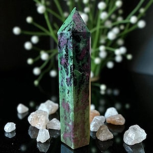 May include: A tall, rectangular crystal point with a green and red mottled pattern. The crystal is surrounded by small, clear and tan-colored stones. The background features white flowers against a dark surface.