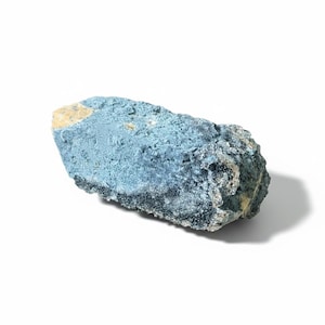 May include: A rough, oblong-shaped rock with a textured surface. The rock is primarily a muted blue color, with areas of black and tan. The rock is set against a white background, casting a shadow.