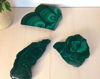 May include: Three polished malachite stone slabs with swirling patterns of dark and light green. The slabs are irregularly shaped and rest on a light wood surface. A woven basket with a green plant is in the background.