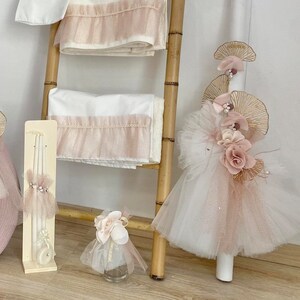 May include: A pink and white baby christening set with a tulle and flower-adorned candle, a pink tote bag, a white towel with pink trim, and a small bottle of holy water.