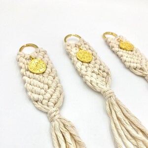 May include: Three white macrame keychains with gold-colored metal rings and charms. The keychains have a tassel at the bottom.