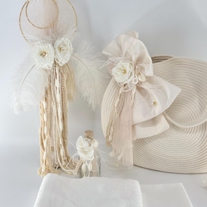 May include: A set of baptism gifts in a neutral colour palette. The set includes a white candle with gold accents, a white and cream woven basket, a white bottle with a white flower, and a white towel with a blush pink lace trim.