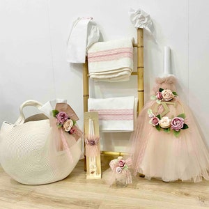 May include: A pink and white baby shower gift basket with a white wicker basket, a pink tulle dress, a white towel, and a white candle with pink flowers.
