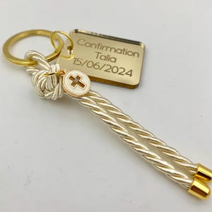 May include: A gold-colored keychain with a rectangular tag engraved with "Confirmation Talia 15/06/2024." The keychain features a white and gold cross charm and a twisted cord in cream and gold. The keychain ring and end caps are gold.